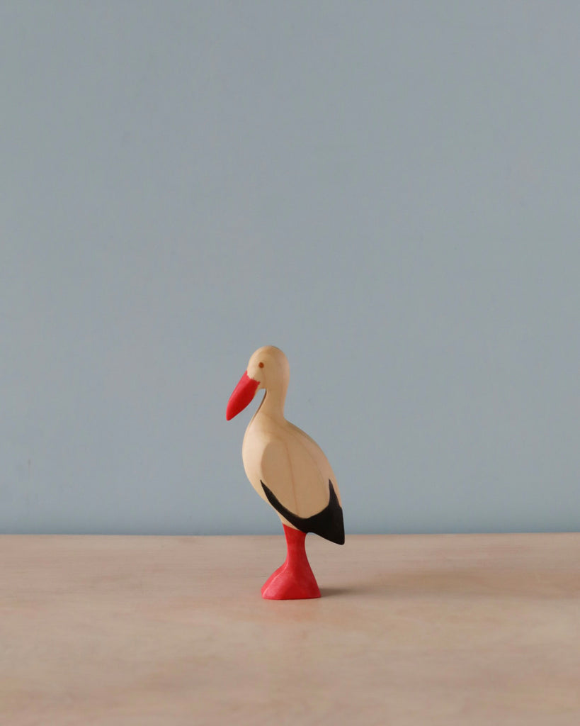 A Handmade Holzwald Stork figurine with a red beak and legs, and a black and white body, standing against a pale blue background.