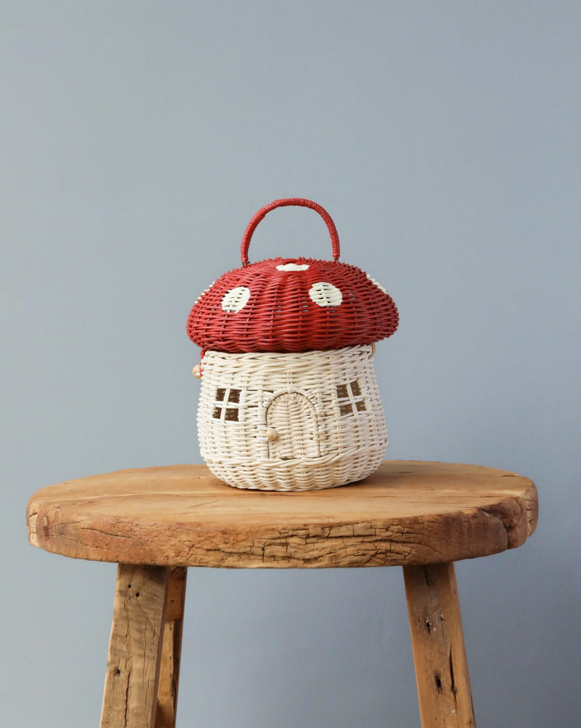 An Olli Ella Red Mushroom Basket, shaped like a mushroom with a red cap and white stalk, placed on a wooden stool against a neutral gray background.