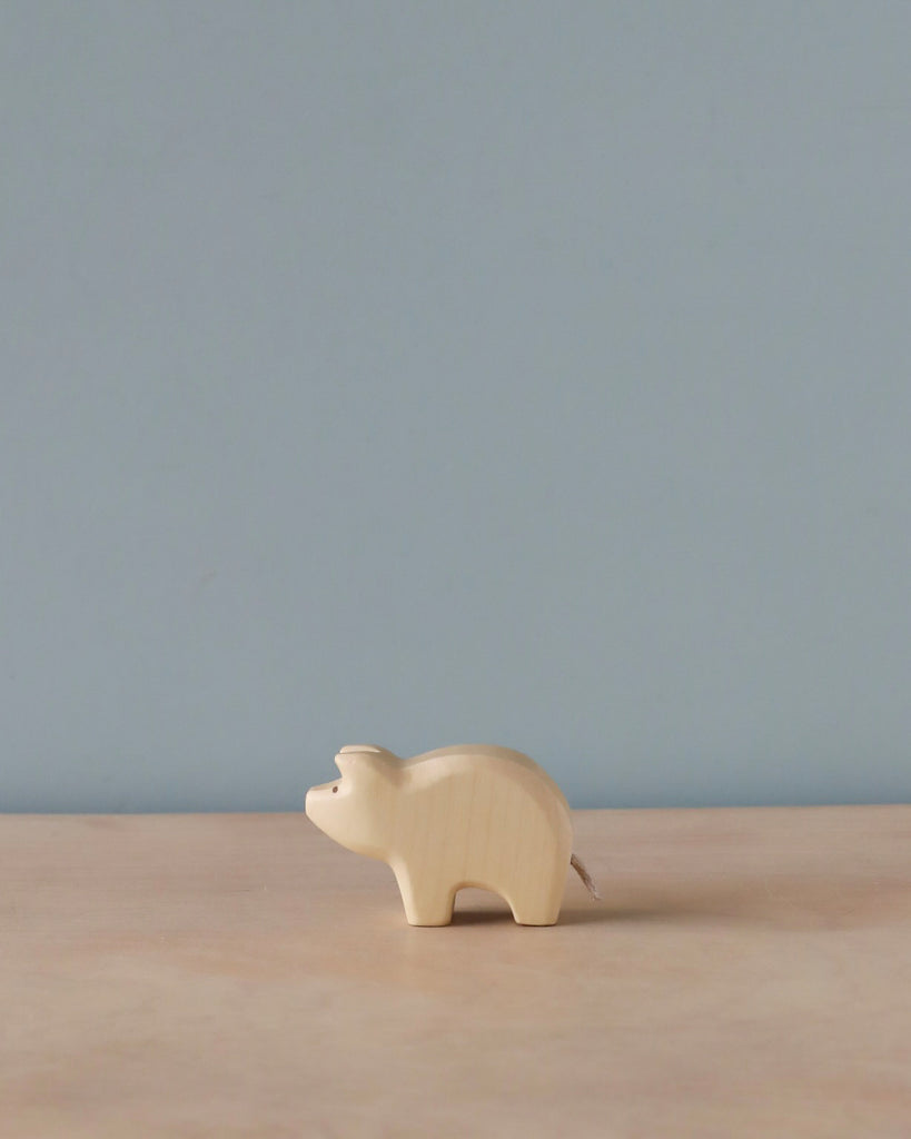 A handmade Holzwald Piglet figurine on a wooden surface against a plain light blue background. The figurine is minimalistic with visible wood grains and represents sustainable toys.