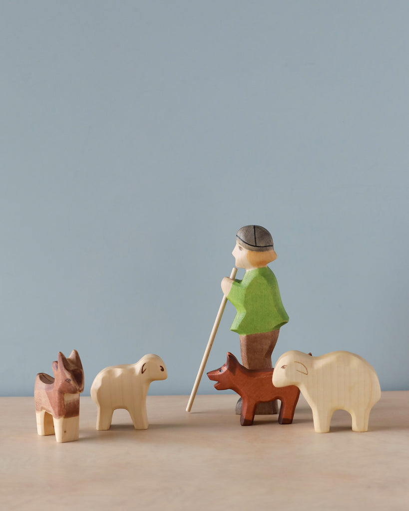 A sustainable Handmade Holzwald Shepherd figurine with a cane, guiding three sheep, each a different color, against a soft blue background.