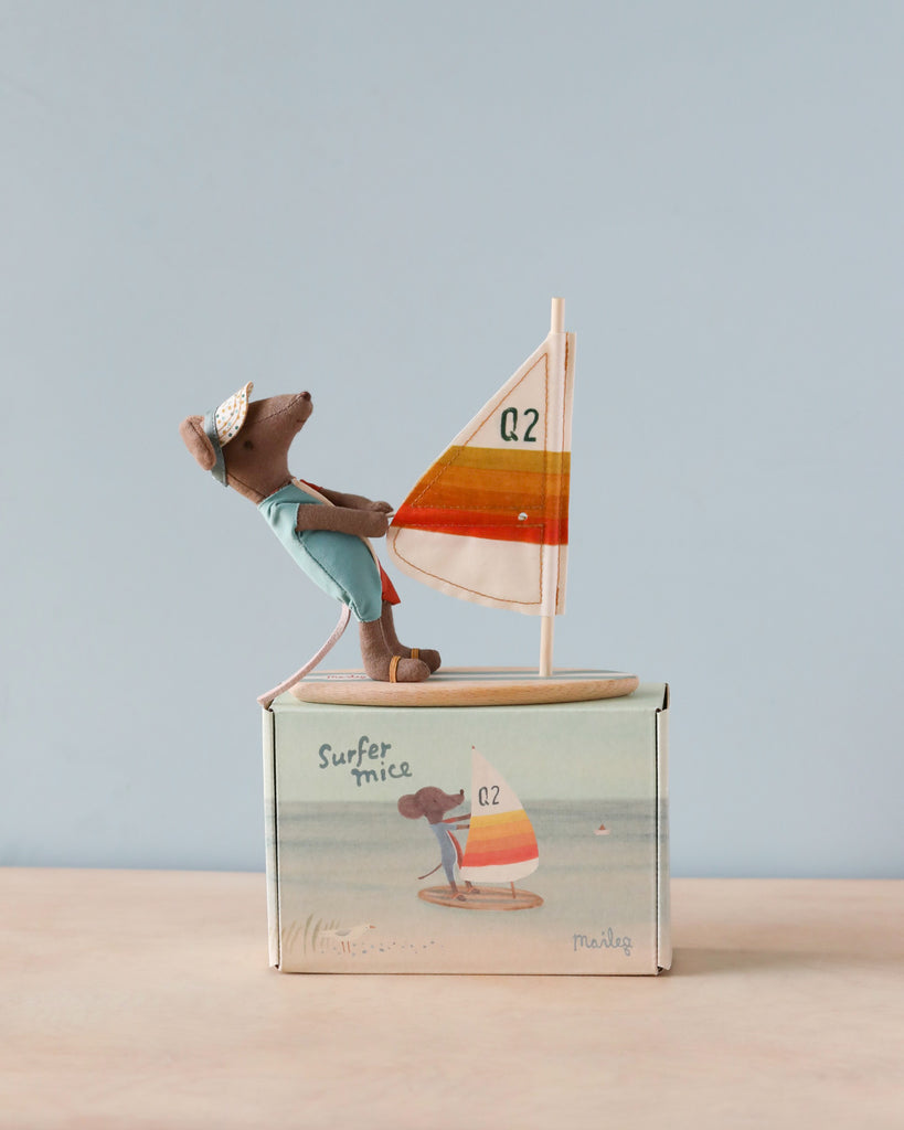 A small, brown toy mouse wearing a light blue outfit and white hat is riding a miniature sailboat with a Q2 sail. The toy is positioned on top of a light blue box that features an illustration of the same mouse sailing. The background, reminiscent of coastal vibes, pairs perfectly with Maileg Surfer Big Brother Mouse adventures.