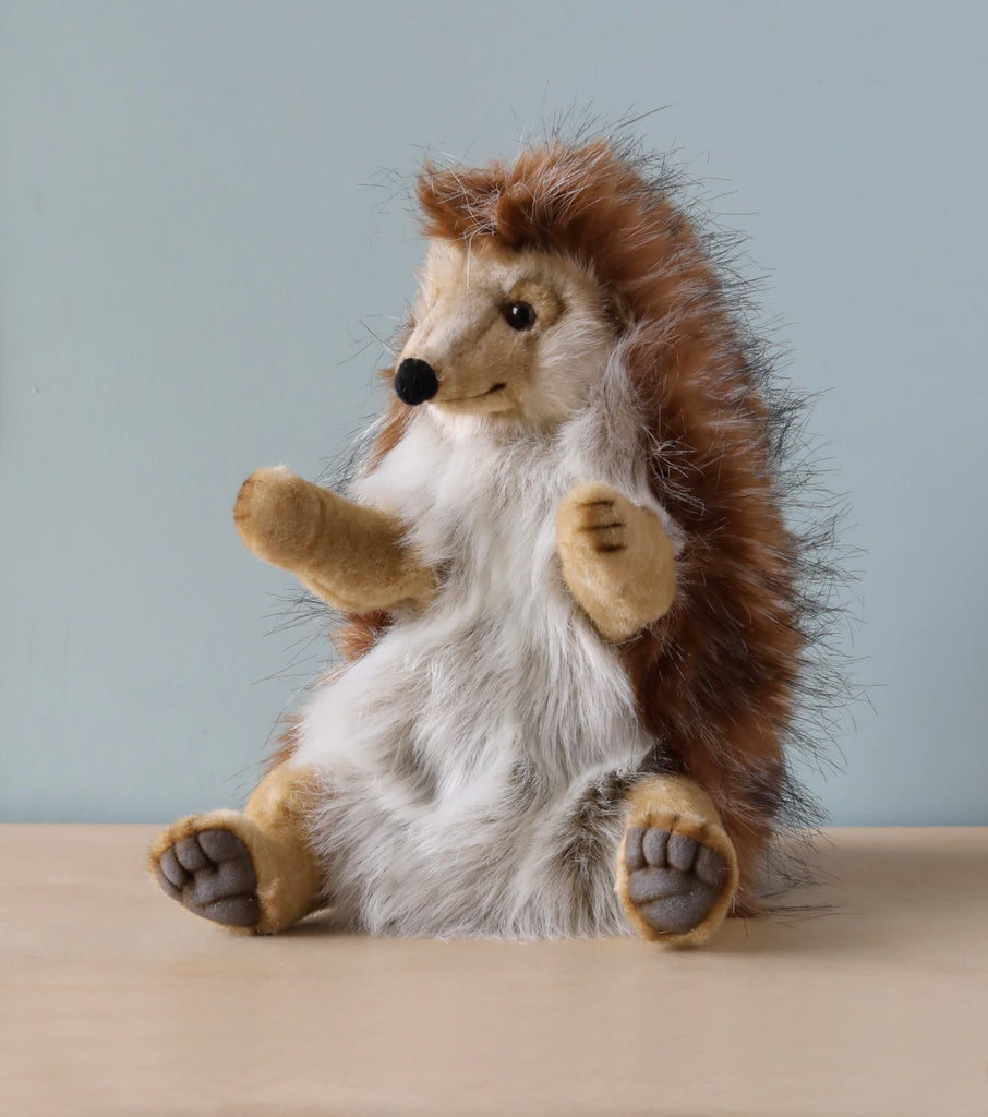 A Hedgehog Puppet with brown fur and a white belly sits upright on a wooden surface against a light gray background. Its small paws and face are detailed, showcasing realistic features that make this hand-sewn plush part of the exquisite HANSA animals collection.