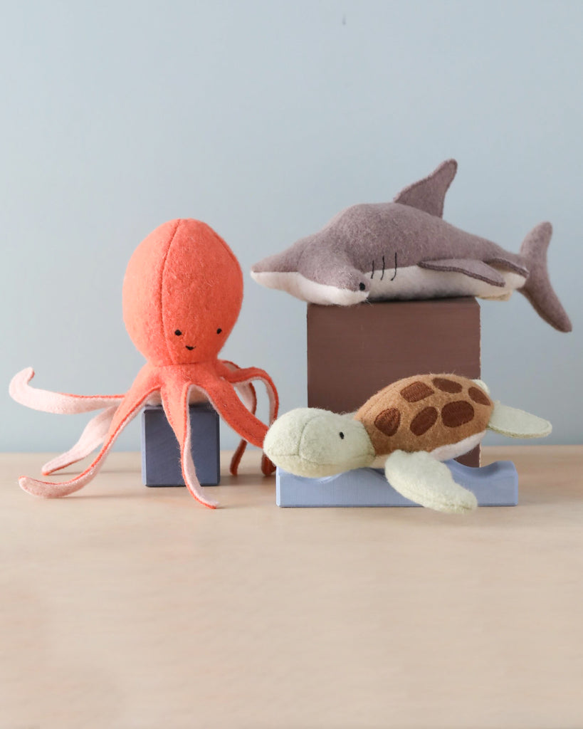 Three Olli Ella Holdie Folk Felt Marine Animals, an orange octopus, a grey shark, and a green sea turtle, are arranged on and around small blocks, set against a plain background.