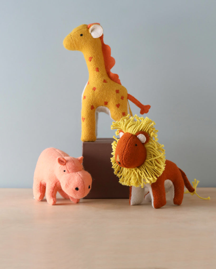 Three Olli Ella Holdie Folk Felt Savannah animals—a giraffe, a lion, and a pig—on a wooden surface against a plain light gray background.