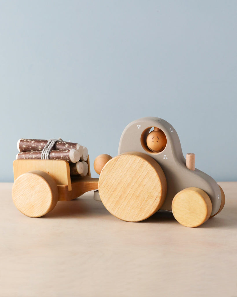 A Handmade Wooden Tractor with large circular wheels is towing a small detachable trailer filled with miniature logs. The toy features a small wooden figure seated inside the tractor. The background is plain light blue, and the scene is set on a light wooden surface.