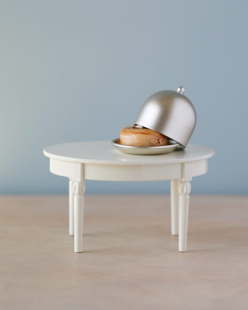 A single burger on a small Maileg Miniature Dining Table with a metal dome cover partially lifted, against a soft blue background.