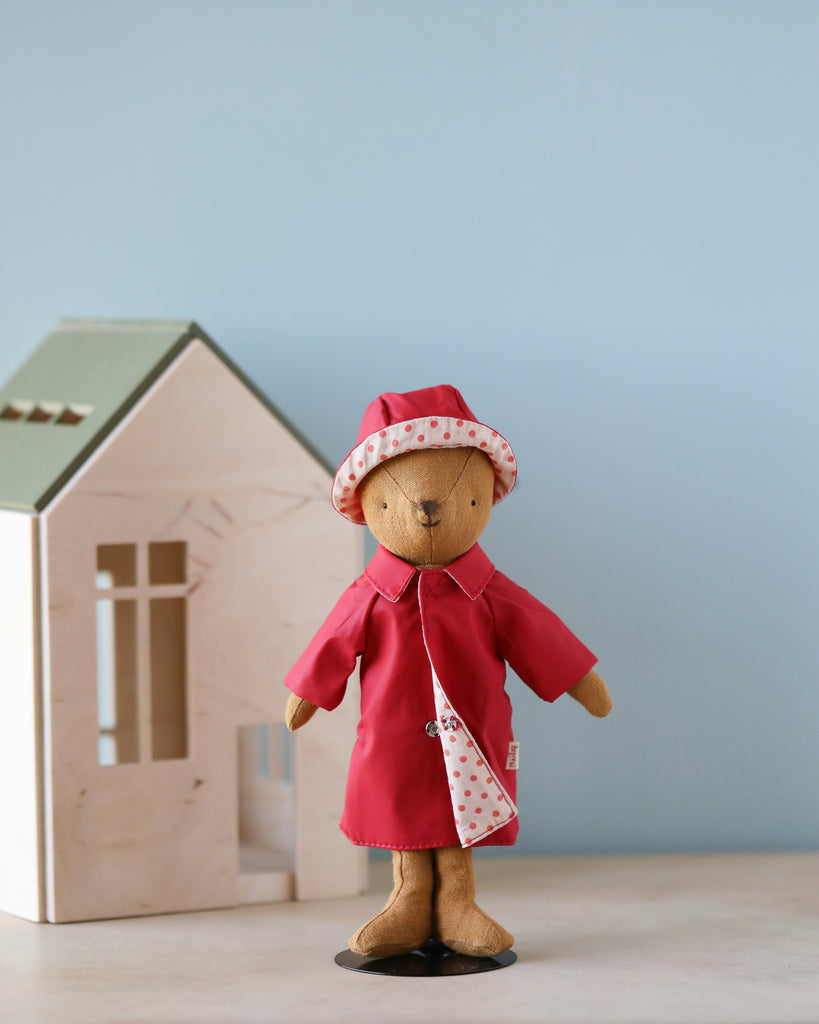 A Teddy Mum in a delightful outfit—Maileg Extra Clothing Rain Coat and matching red hat with polka dot trim—stands in front of a small wooden house. The light blue background adds a soft and playful atmosphere to the scene.