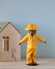 A stuffed animal, lovingly referred to as Teddy Dad, is dressed in the delightful Maileg Extra Clothing: Rainwear for Teddy Dad, featuring a charming yellow raincoat and hat. It stands on a surface beside a small wooden toy house against a plain blue wall, proudly displaying its adorable rainwear outfit.