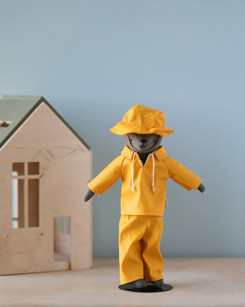 A stuffed animal, lovingly referred to as Teddy Dad, is dressed in the delightful Maileg Extra Clothing: Rainwear for Teddy Dad, featuring a charming yellow raincoat and hat. It stands on a surface beside a small wooden toy house against a plain blue wall, proudly displaying its adorable rainwear outfit.