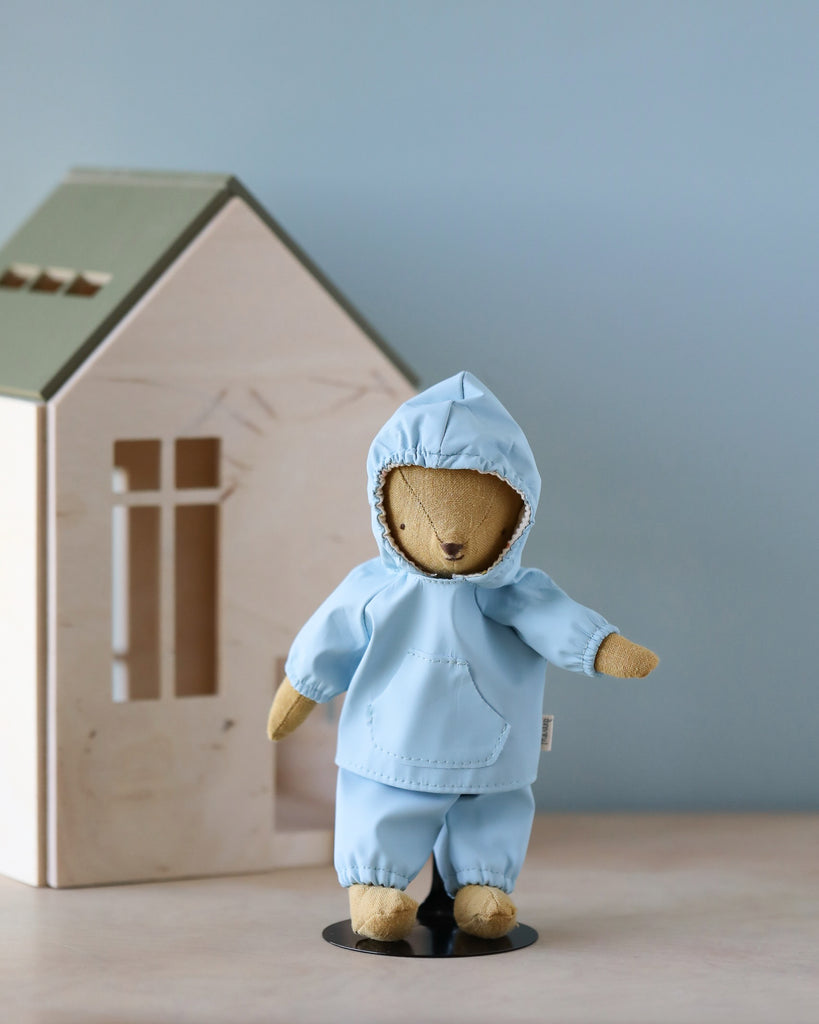 Teddy Junior, looking charming in the Maileg Extra Clothing rainwear outfit—a delightful ensemble of a light blue raincoat and pants—stands proudly in front of a small wooden model house with a green roof. The light blue backdrop further enhances the serene and playful mood.