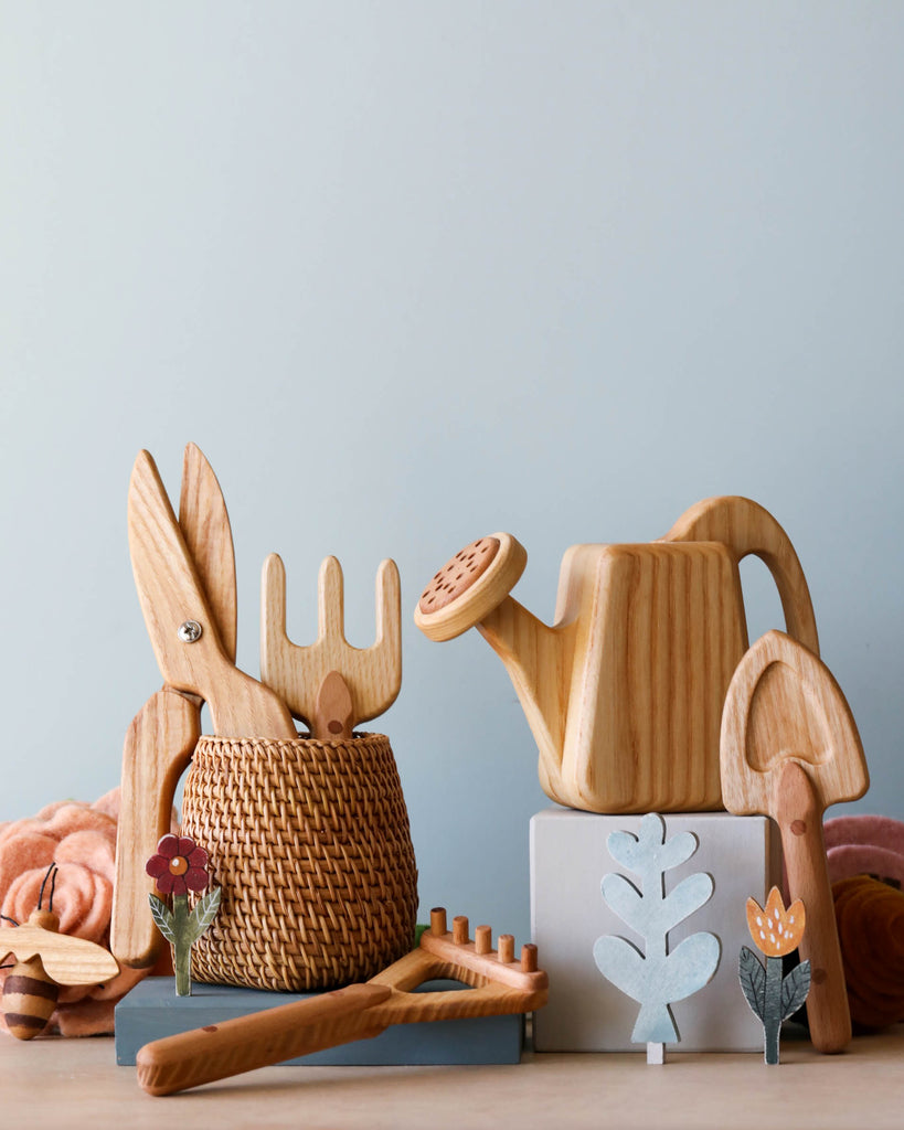 A delightful assortment from the "Handmade Pretend Play Gardening Tools" collection is neatly arranged on a surface, featuring wooden garden tools such as a shovel, rake, and watering can. Complementing the set are wooden plant decorations and a wicker basket. The backdrop of plain light blue enhances the scene’s minimalist charm.