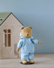 A teddy bear named Teddy Junior, dressed in light blue pajamas and a bonnet, is positioned beside a wooden toy house against a soft blue background.