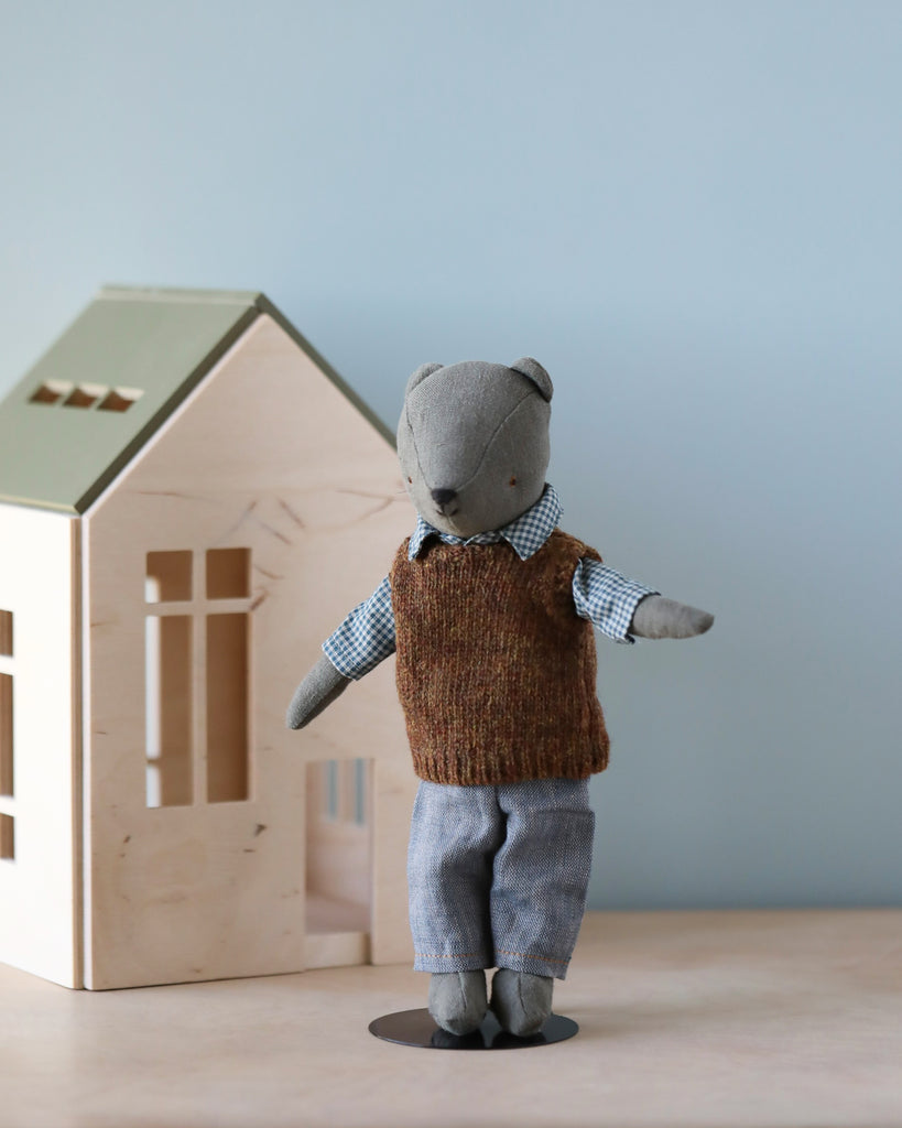 A small, plush toy bear dressed in a brown knitted vest, blue and white plaid shirt, and blue pants stands upright in front of a light wood toy house with green roof accents. Named "Teddy Dad," his stylish outfit, Maileg Extra Clothing: Shirt, Slipover and Pants for Teddy Dad, can be purchased separately. The background is a plain, light blue wall.