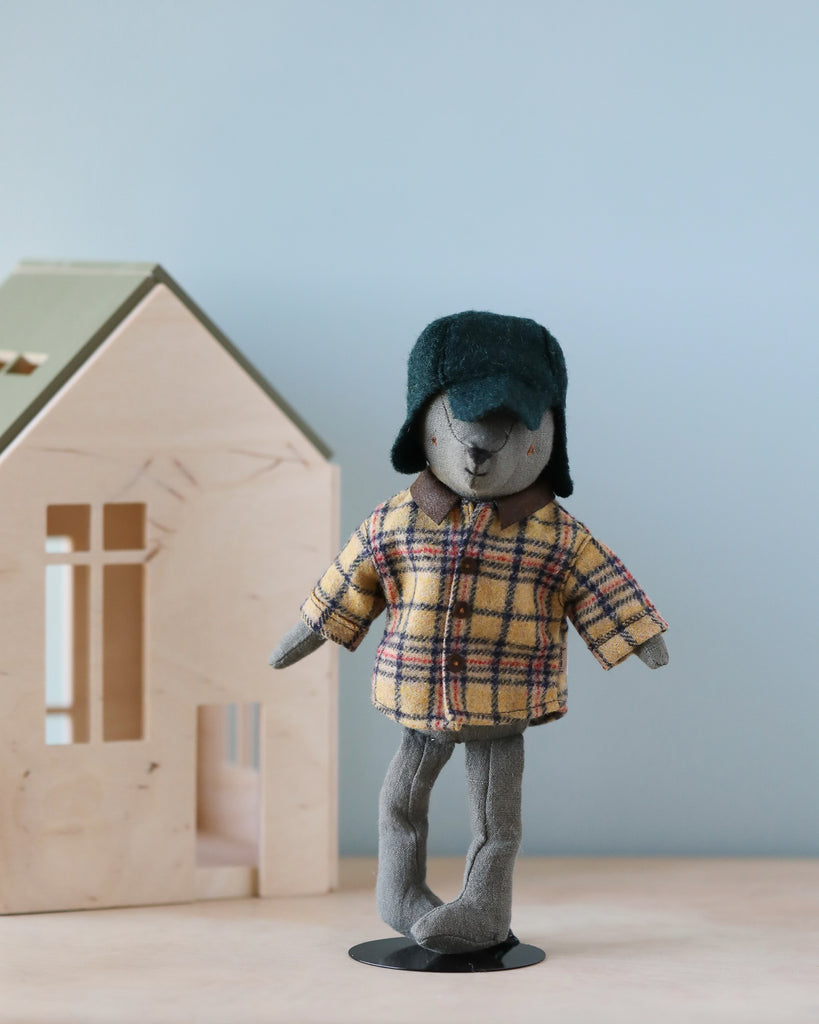 A plush Teddy Dad, adorned in a "Woodsman Jacket and Hat" ensemble, stands upright on a light wood surface. Behind him, a wooden model of a house rests against a light blue background. This stylish outfit, known as the Maileg Extra Clothing: Woodsman Jacket and Hat for Teddy Dad, is available for purchase separately to enhance customization options.