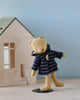 A stuffed teddy bear wearing a blue and white striped Teddy Junior outfit and a Maileg Duffle Coat for Teddy Junior stands on one leg on a light wooden surface. Behind the bear is a small wooden house with a mint green roof, set against a light blue background.