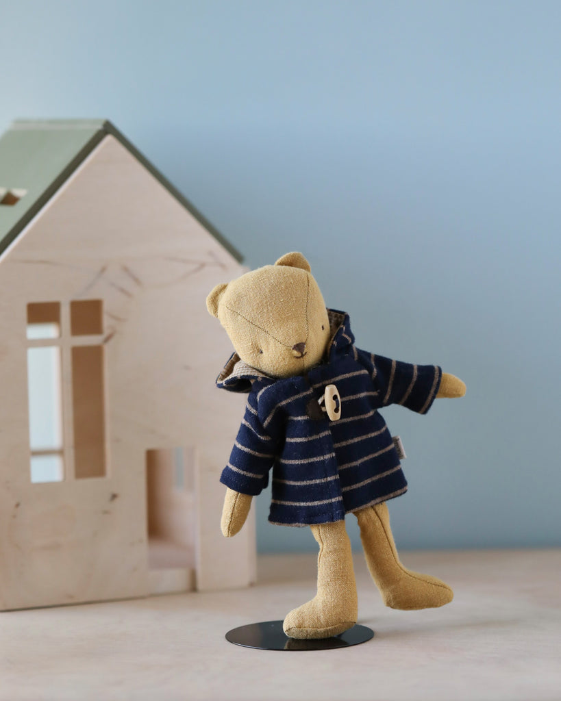 A stuffed teddy bear wearing a blue and white striped Teddy Junior outfit and a Maileg Duffle Coat for Teddy Junior stands on one leg on a light wooden surface. Behind the bear is a small wooden house with a mint green roof, set against a light blue background.