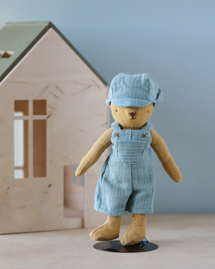 A small stuffed bear named Teddy Junior stands upright in front of a wooden toy house. The bear is dressed in light blue overalls and a matching cap, both made of striped fabric, which you can purchase separately as the Maileg Extra Clothing: Overall and Cap for Teddy Junior. The background is a soft blue color, creating a calm and inviting scene.