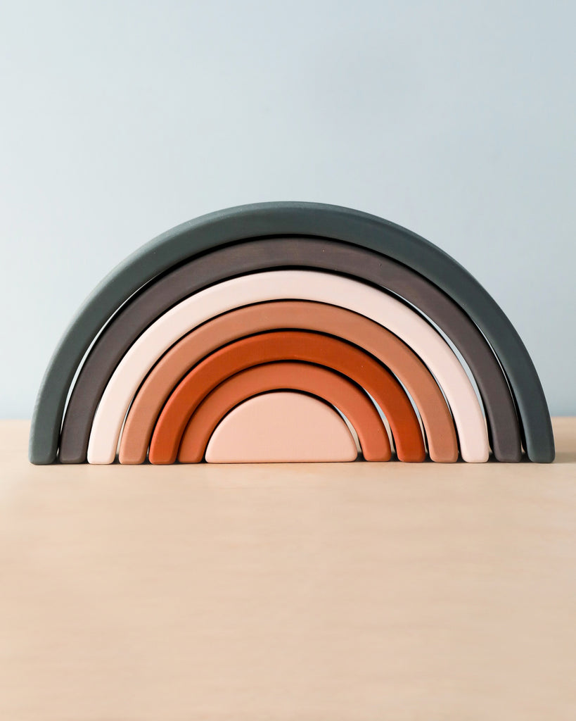 A neatly arranged stack of semi-circular, rainbow-shaped wooden pieces sits on a light wooden surface. The Handmade Rainbow Stacker - Mojave Desert features pieces that vary in size and are colored in shades of gray, brown, and pink using non-toxic paint.