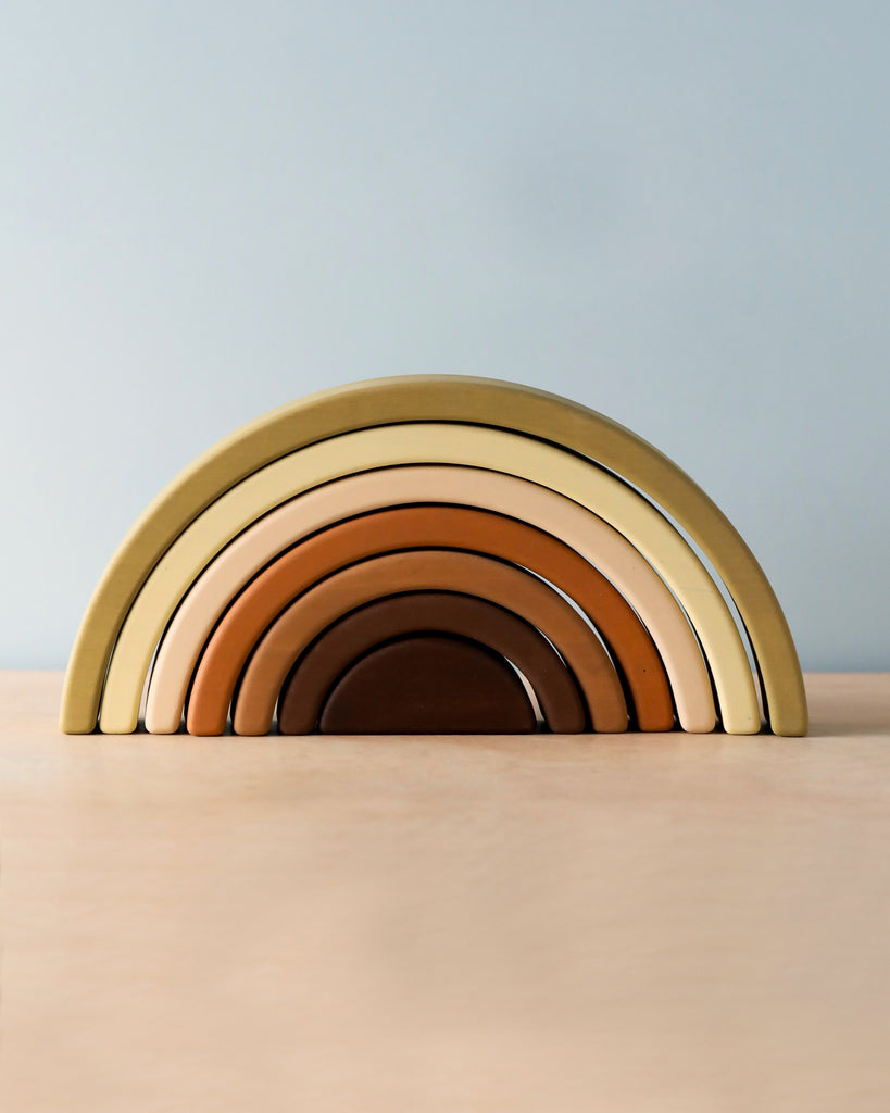 A Handmade Rainbow Stacker - Olive with seven arches, each painted in shades of brown, beige, and mustard yellow using non-toxic paint, is displayed on a light wooden surface against a soft blue background.