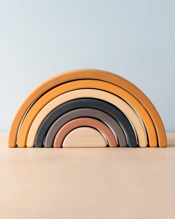 The Handmade Rainbow Stacker - Mustard is a minimalist, non-toxic wooden rainbow stacker toy featuring seven handmade arches in neutral shades such as beige, brown, gray, and taupe. These arches are artfully arranged on a light wooden surface, set against a plain light blue background.