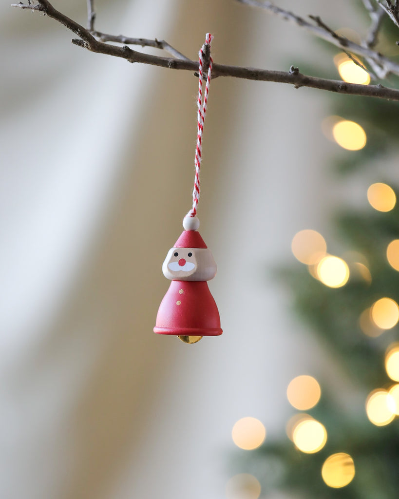 Close-up of a Christmas ornament hanging on a tree branch. The 2 x 1.5 inches Wooden Santa Ornament, shaped like a small figure with a red hat, white beard, and red coat resembling Santa Claus, includes a decorative bell. The background features blurred Christmas lights and greenery.