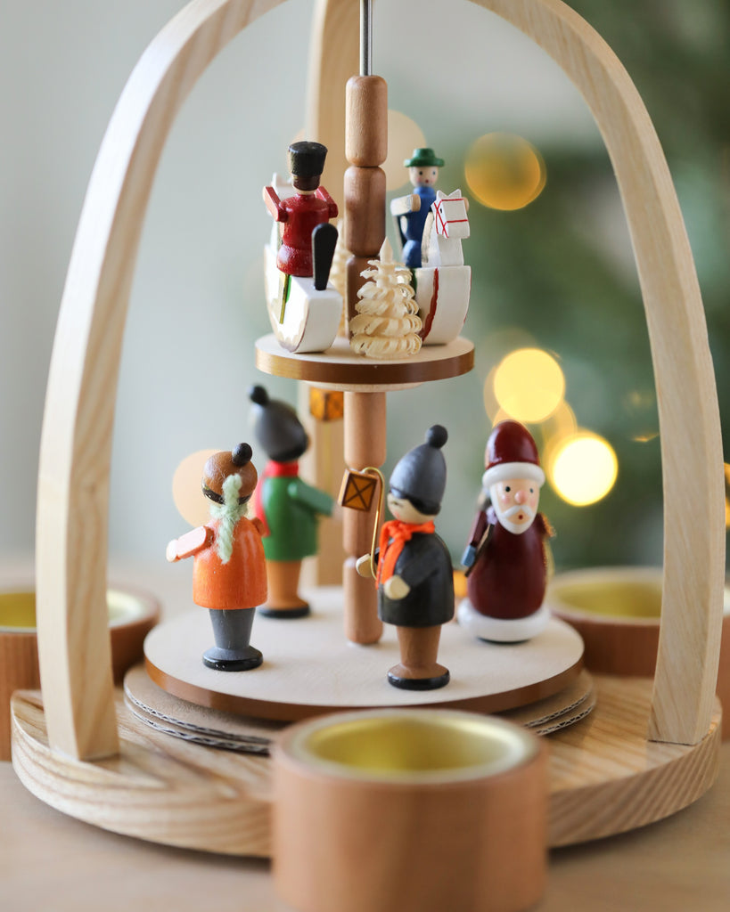 A Christian Ulbricht Two Tier Christmas Pyramid with hand-painted wooden figures, including a Santa Claus, arranged on rotating tiers set against a softly blurred holiday backdrop.