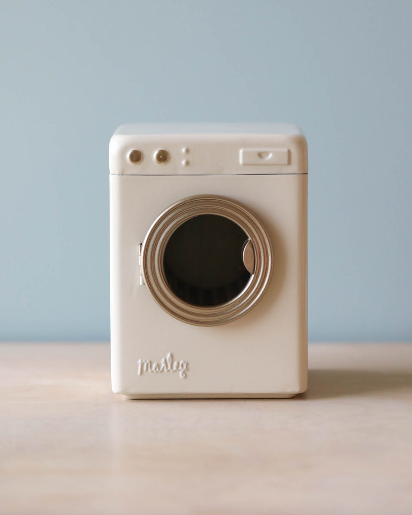 A vintage white Maileg Washing Machine with a prominent lens and flash, positioned upright on a wooden surface against a light blue background.