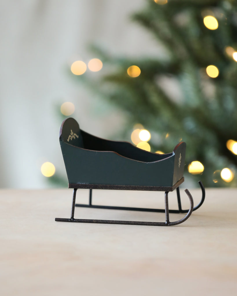 A Maileg Christmas Sleigh - Green with black runners and a dark green body is placed on a flat surface. Hand-painted details add charm, while the background features a blurred, lit Christmas tree with yellow lights, giving a festive ambience.