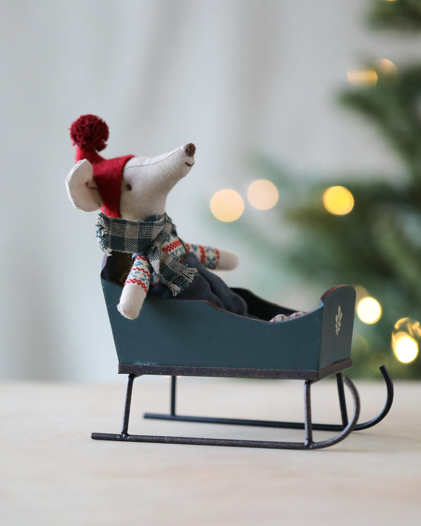 A small plush mouse wearing a red hat and a patterned sweater sits in a Maileg Christmas Sleigh - Green. The background shows blurred Christmas lights and part of a tree, creating a festive atmosphere with intricate hand-painted details.