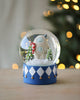 The Yeti Snow Globe features a charming yeti with a candy cane amidst small snow-covered Christmas trees, set against a softly blurred backdrop illuminated with warm lights. The blue base is adorned with a harlequin pattern.