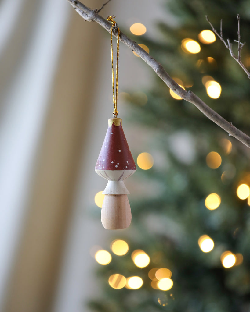 The Handmade Wooden Mushroom Ornament, featuring a red cap with white spots and standing at 4 inches tall, hangs gracefully from a branch. Against the backdrop of a blurred Christmas tree adorned with warm white lights, it creates a festive atmosphere and stands out beautifully.