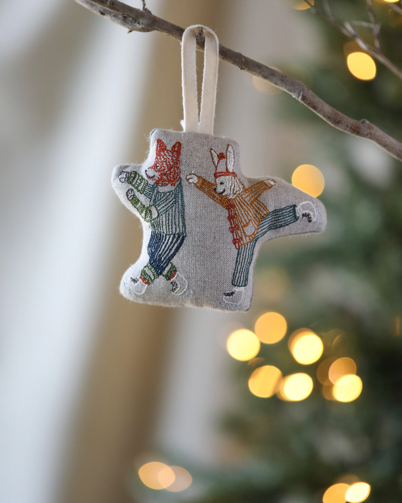 Coral & Tusk Ice Skater Bunny and Fox Ornament featuring two foxes dressed in figure skating outfits, ice skating, hangs from a Christmas tree, with softly blurred lights in the background.