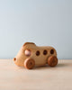 A handmade wooden bus with a simplistic design, featuring round holes as windows and circular, darker wooden wheels, set against a plain light blue backdrop.