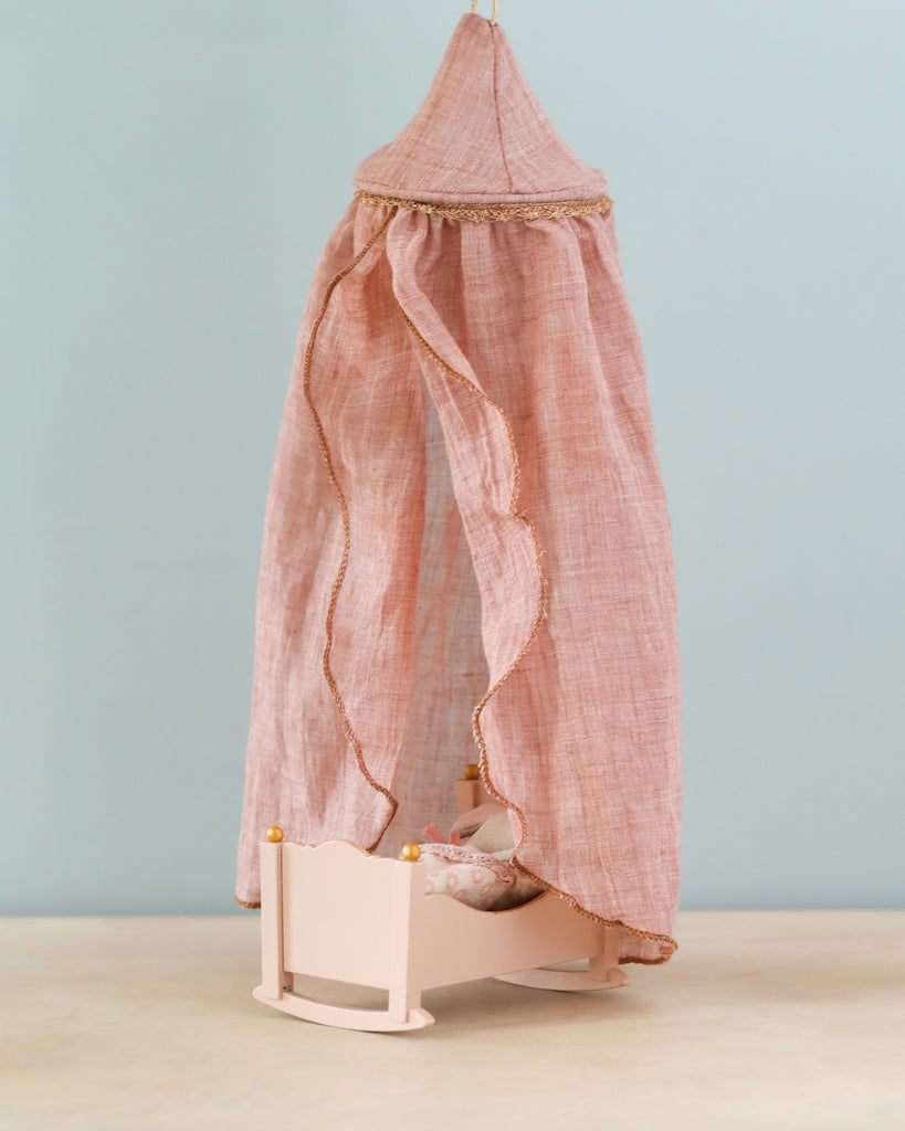 A Maileg Mini Bed Canopy with a small doll lying in it, covered by a pink blanket. A pink and gold canopy drapes over the bed, adding to the cozy and elegant atmosphere. The bedroom ceiling is adorned with delicate gold accents that complement the bed's decorations. The background is a simple light blue wall.