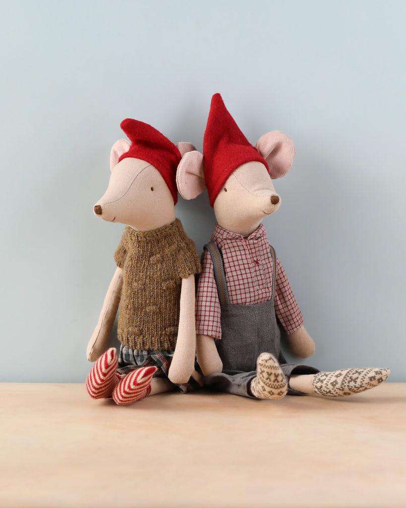 Two Maileg Medium Christmas Mice sit side by side against a light blue wall. Both Christmas mice wear red hats. The left mouse wears a brown knitted sweater, plaid pants, and striped socks, embodying cozy clothes. The right mouse is dressed in a checkered shirt, grey dungarees, and socks with a geometric pattern.