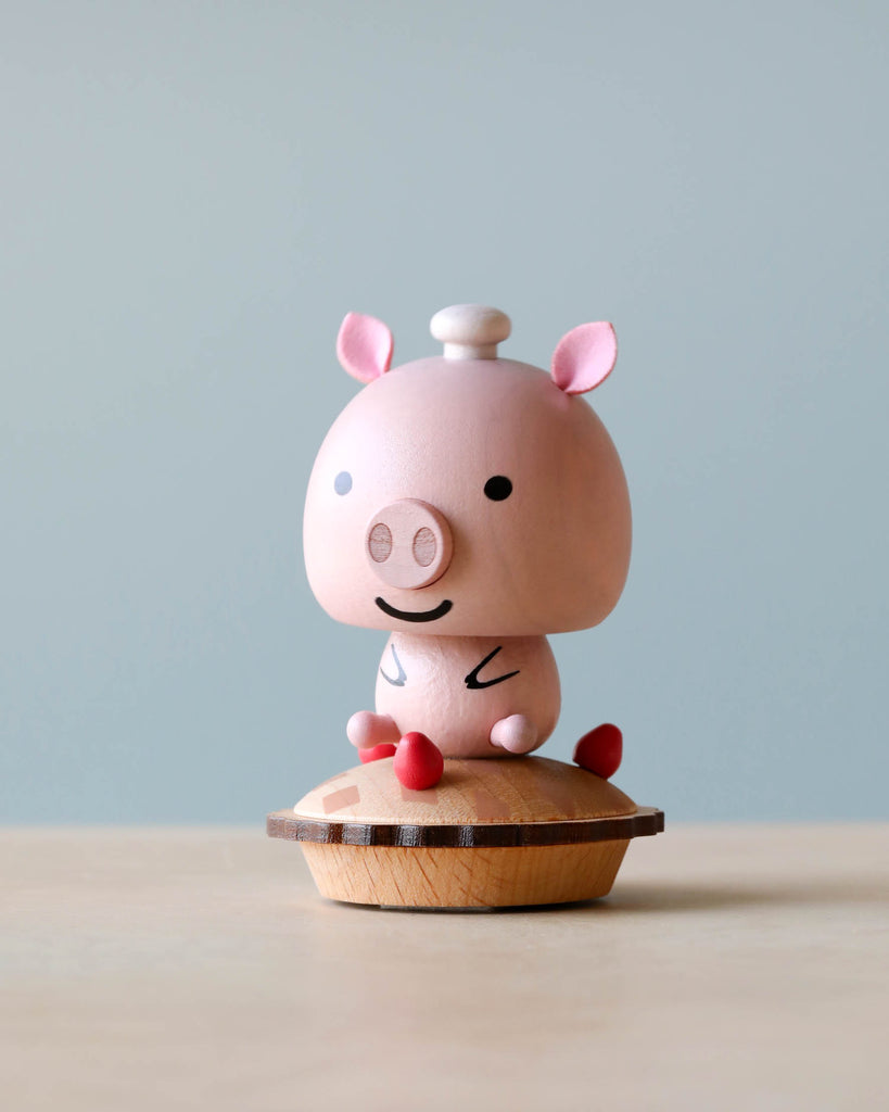 A Wooden Pig Bobblehead with a smiling face, standing on a small wooden stand. The pig is light pink with darker pink ears, a tiny lid on top of its head.