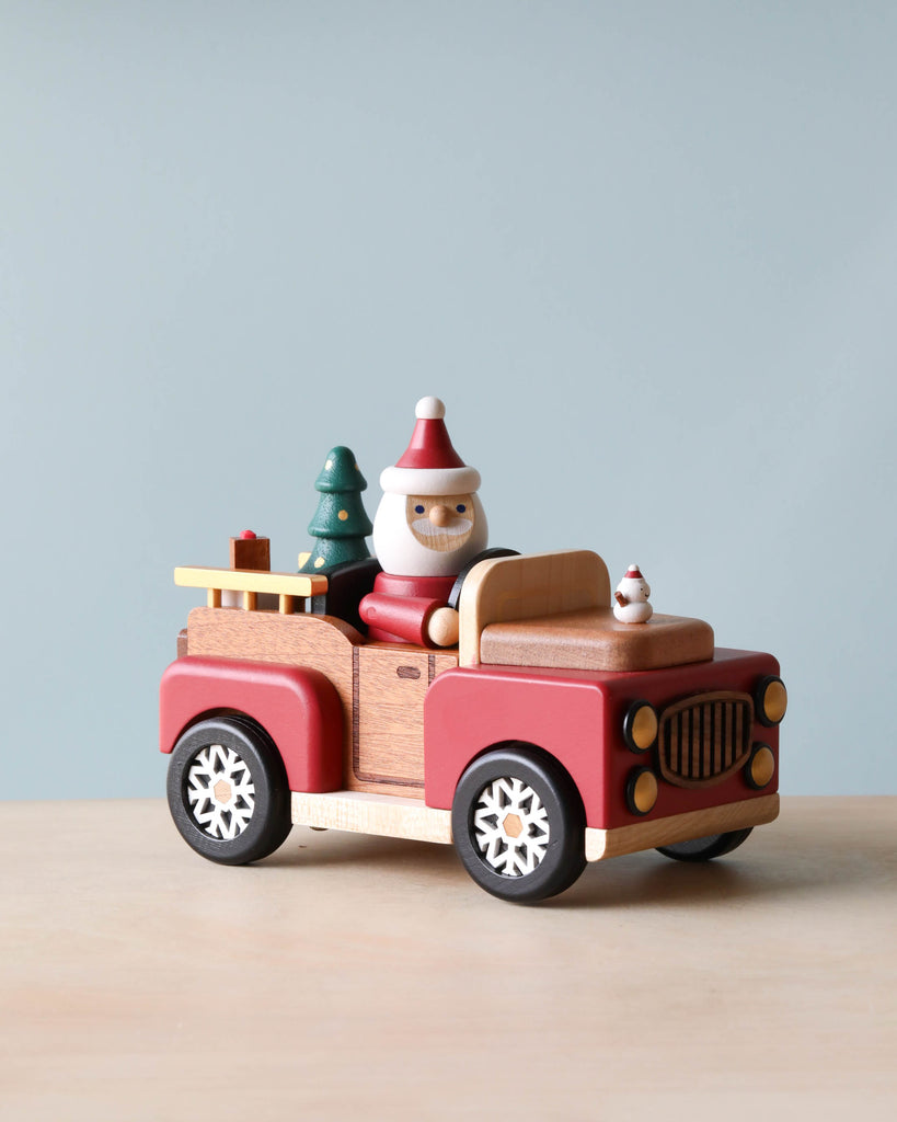 A Santa-mobile Music Box, crafted from premium hardwoods and painted red and beige with white wheels, carries charming wooden figures: a snowman driver, a small Christmas tree, and a puppy in the back.