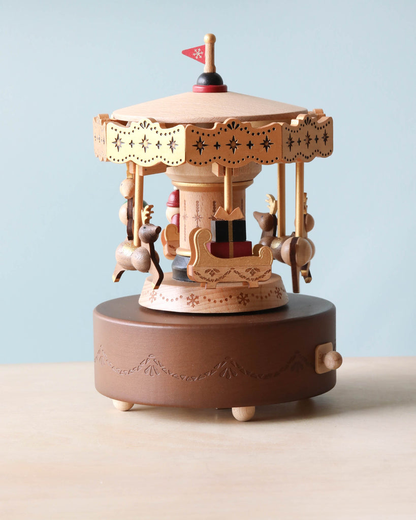 Christmas Carousel Music Box featuring reindeer and a sleigh, intricately carved with delicate patterns from sustainably sourced wood. The hand-cranked music box sits atop a round base with a side crank handle, designed for ages 12 and up, against a light blue background.