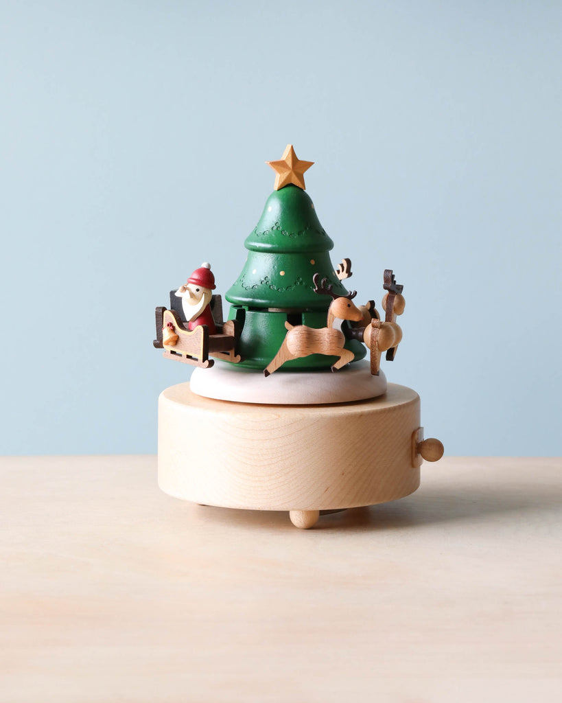 The Santa & Reindeer Music Box, hand-cranked and made from sustainably sourced wood, features a charming green Christmas tree topped with a yellow star. Encircling the base are intricately carved figures of Santa Claus and reindeer, offering an ideal decorative touch against a simple light blue background.