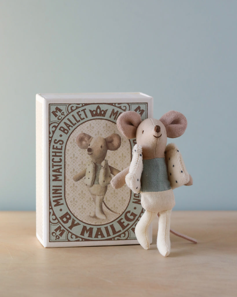 The Maileg Little Brother Dancer Mouse in a Box, dressed in a charming ballerina outfit, stands proudly in front of its matchbox-style packaging. The vintage-themed box features an illustration of another dancing mouse. This delightful character boasts beige ears, a blue top, white pants, and an eye-catching cape.