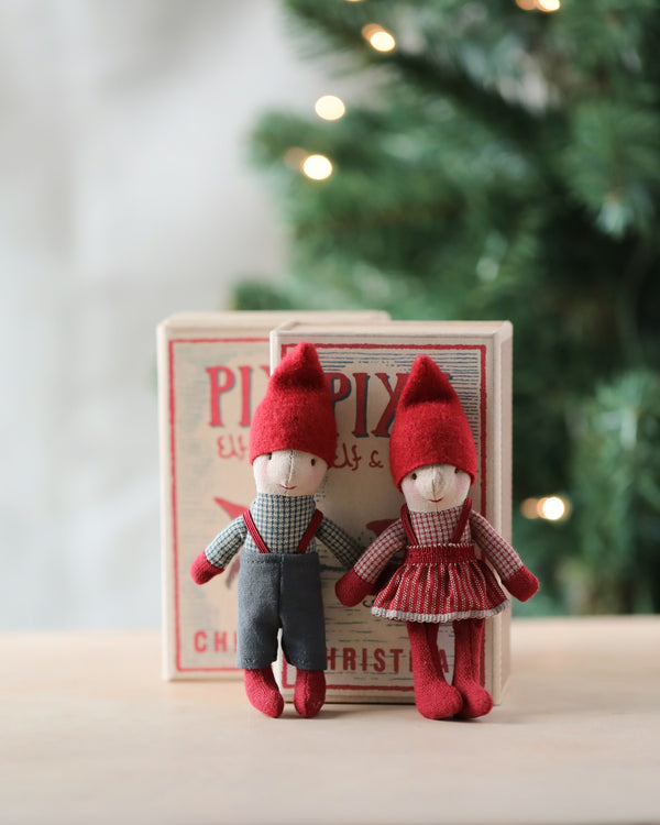 Two small elf dolls with red hats, standing in front of a festive box labeled "Maileg Pixy Elfie." The elf on the left, your new Christmas friend, wears gray pants and a checkered shirt, while Maileg Pixy Elfie on the right wears a red dress. A blurred Christmas tree with lights shines in the background.