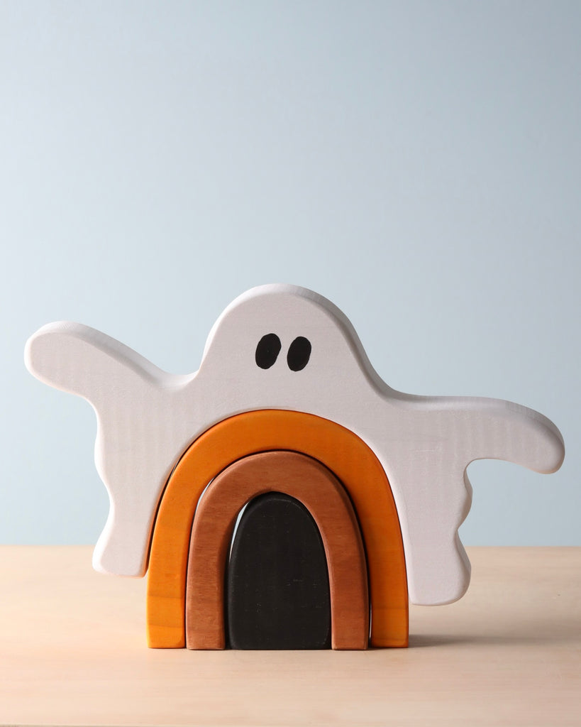 A Handmade Wooden Ghost Stacker in the shape of a ghost with arms outstretched. The ghost, crafted from poplar hardwood and adorned with black eyes, is stacked above curved pieces in brown and orange tones. Non-toxic wood dye enhances its charm, all set against a light blue background on a wooden surface.