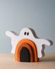 A Handmade Wooden Ghost Stacker, crafted from poplar hardwood with arms outstretched, features black eyes and a mouth. Below the ghost are three arch-shaped pieces in black, orange, and white using non-toxic wood dye. The background is a plain light blue wall.