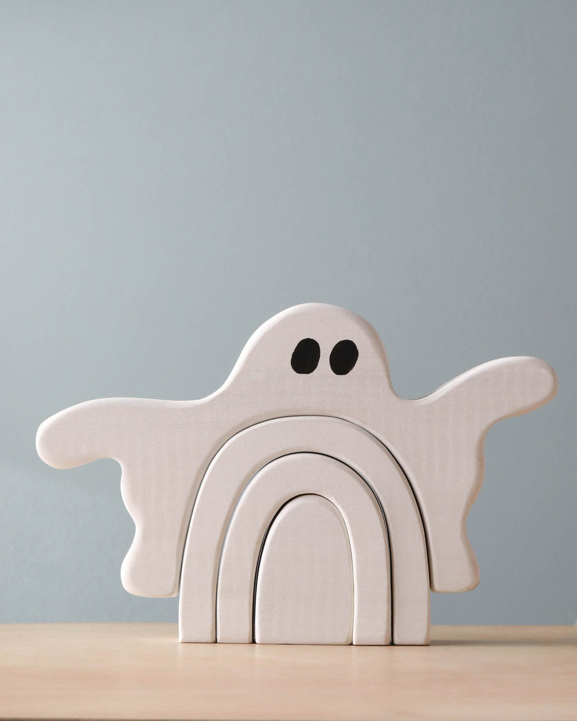 A Handmade Wooden Ghost Stacker, shaped like a ghost with outstretched arms and black eyes, handcrafted from poplar hardwood and finished with non-toxic wood dye, consists of three interlocking pieces. It is placed on a light wood surface against a light gray background.