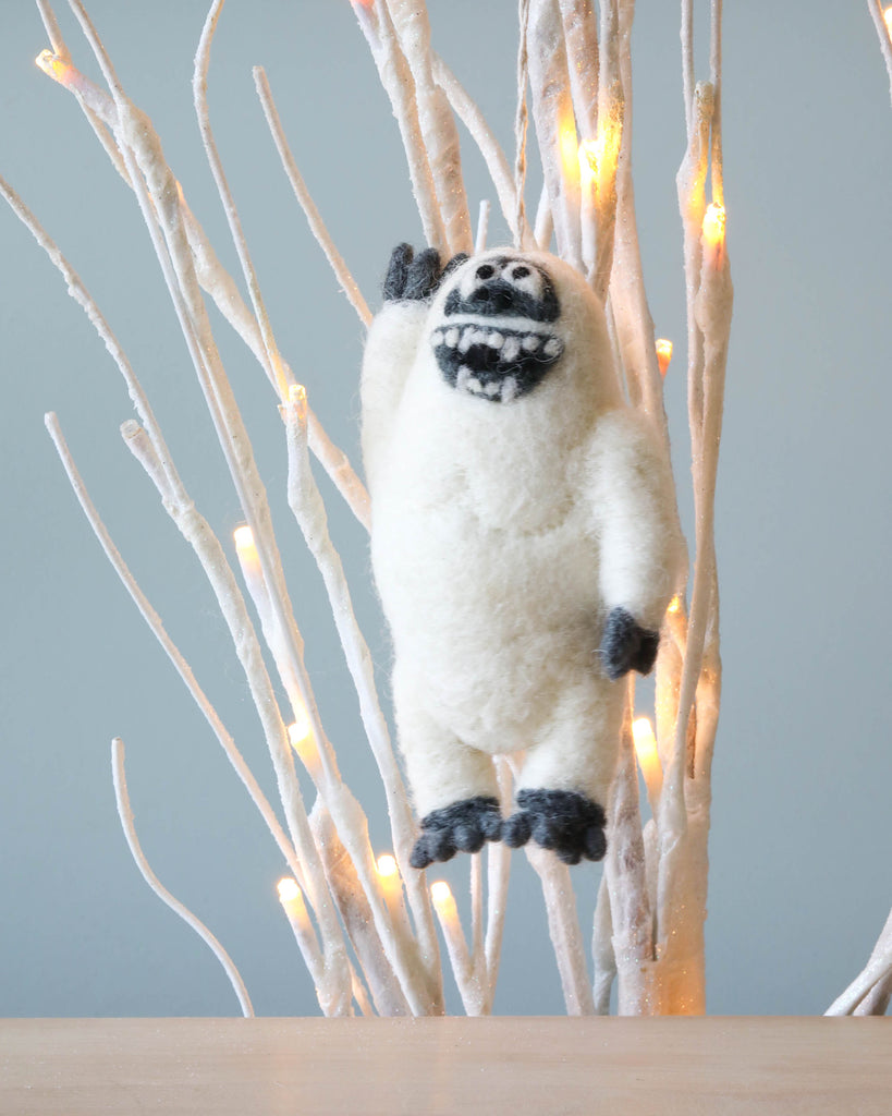 A plush toy character resembling a friendly Handmade Felt Yeti Christmas Tree Ornament, crafted in white and black colors, hanging among white twig-like branches adorned with soft glowing lights against a gray backdrop.