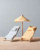 Two Maileg Beach Chairs And/Or Umbrella, one blue and white and the other yellow and white, sit side by side under a matching striped beach umbrella with a wooden stand. The setup is placed on a light wooden surface against a plain gray background.