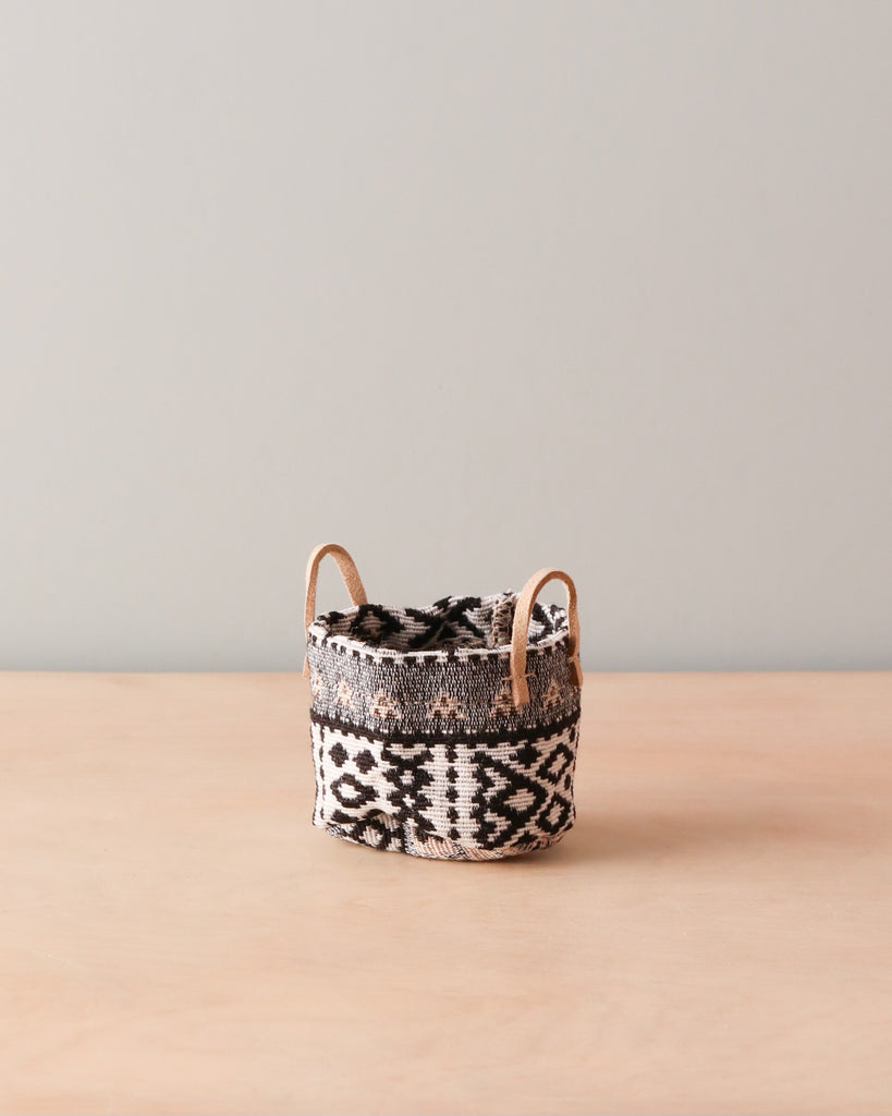 A Maileg | Miniature Basket with geometric patterns and leather handles on a wooden surface against a neutral background.