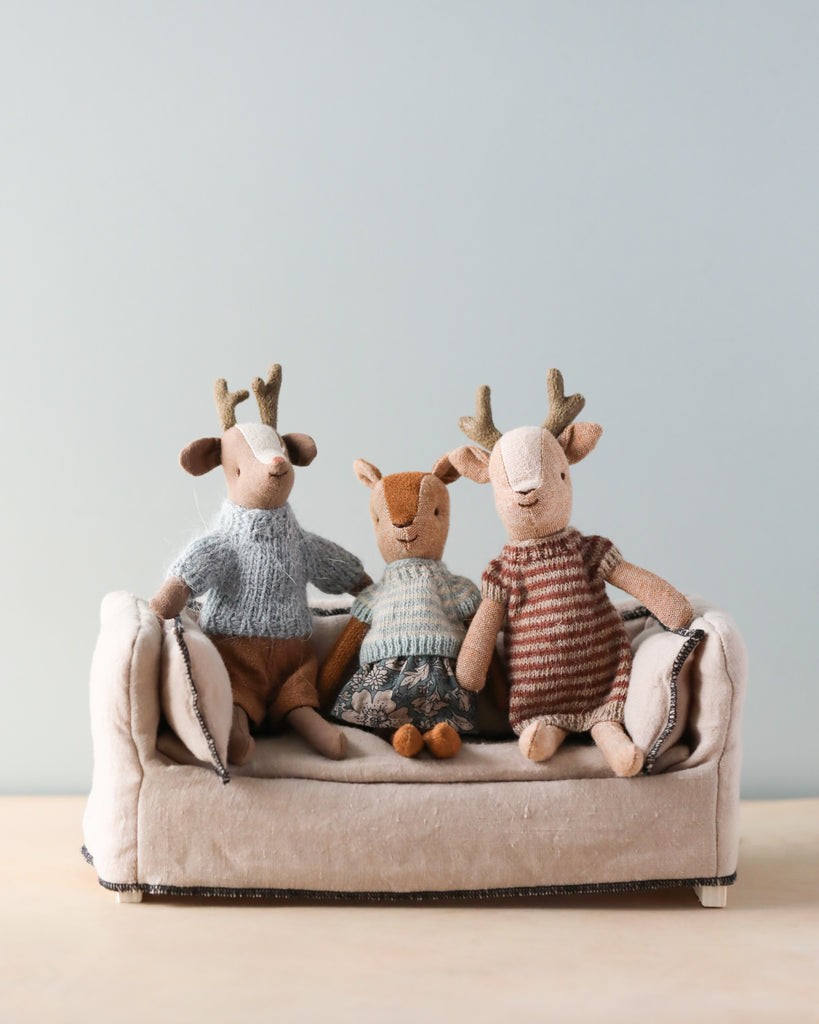 Three plush deer toys are sitting on a small beige sofa. The deer on the left is wearing a blue sweater, the one in the middle is in a Maileg Knitted Dress For Mum Mouse, and the deer on the right is wearing a brown striped shirt. The background is a plain light blue wall.