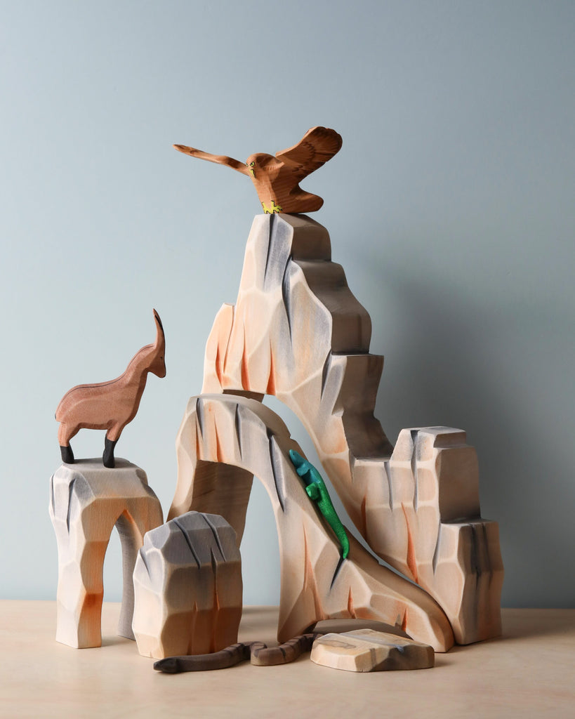 A Handmade Wooden Mountain Cliff Scene. The Waldorf inspired play space includes a rugged mountain, an eagle perched at the peak, a mountain goat on a ledge, and a green lizard climbing up the side—all figures carved with simple outlines and painted with non-toxic paints.