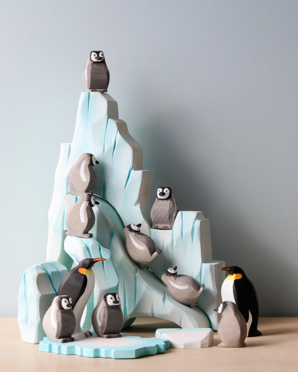 The Handmade Wooden Penguin Set, varying in size, is arranged on and around a stylized wooden iceberg structure with a light blue and white color scheme. Handmade with non-toxic paints, the penguins boast minimalist features in shades of gray, black, and white, creating a charming Waldorf inspired play scene.